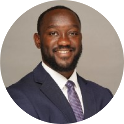 Professional headshot of a man wearing a dark navy suit jacket and purple patterned tie against a neutral gray background.