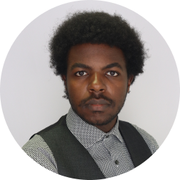 Portrait of a man with dark hair wearing a dark vest over a checkered shirt against a light gray background.