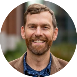 Headshot of a bearded man with reddish-brown hair wearing a blue patterned shirt and brown jacket, smiling at the camera.