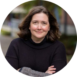 Woman with shoulder-length dark wavy hair wearing a black hoodie, smiling at the camera outdoors on a university campus.
