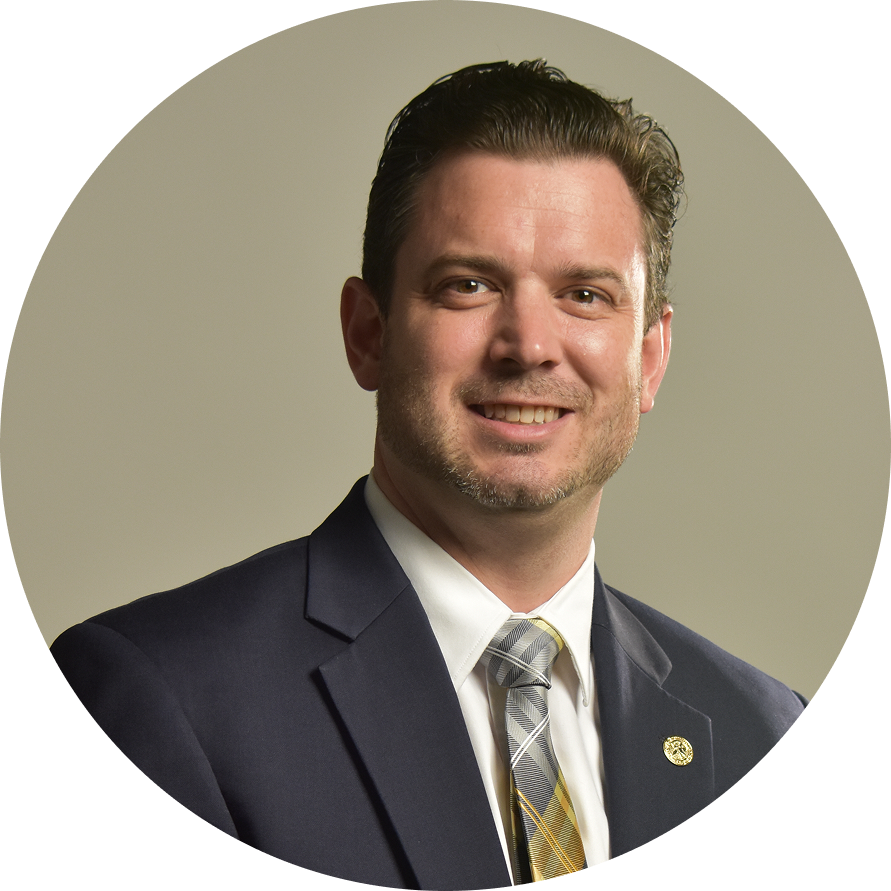 Professional headshot of Dr. Nick Vick wearing a dark blazer, white dress shirt, and patterned tie with a gold lapel pin against a neutral tan background.