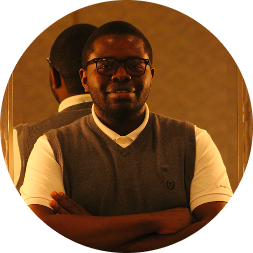 Portrait photograph of a man wearing glasses, a brown vest, and tan and white striped shirt against a warm brown background.