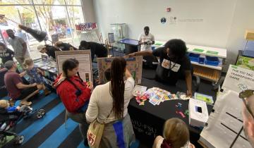 University researchers demonstrate interactive STEM projects to visitors at an indoor outreach event, with children engaging with educational games and displays on tables.
