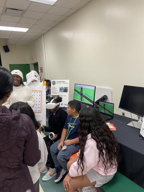 Students gather around a computer display showing a transportation research simulation, with a presenter in white attire demonstrating the interactive project.