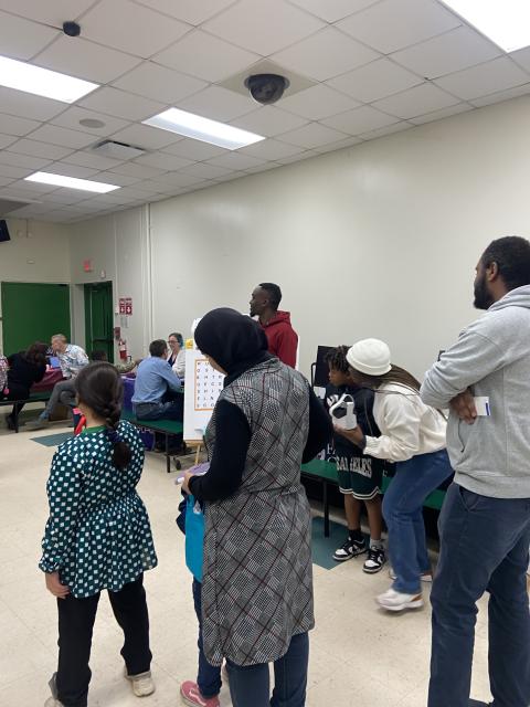 Community members and researchers gather in a classroom for a health screening or educational event, with participants standing and seated around tables.
