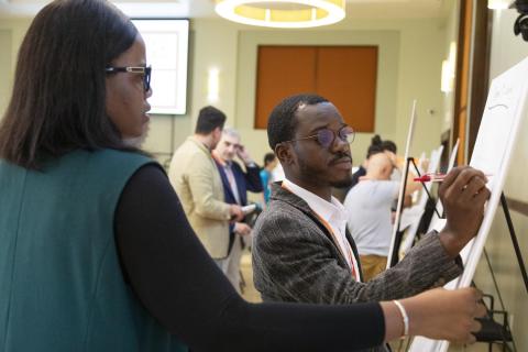 Man wearing glasses and lanyard writing on whiteboard while woman in teal jacket observes during indoor workshop or conference event.