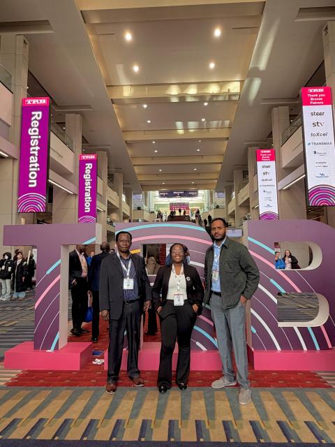 Three attendees wearing conference badges stand in front of a large purple registration sign at a convention center entrance.