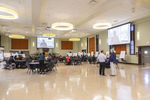 A spacious conference room with attendees seated at round tables and standing in small groups, with presentation screens displaying video content and poster boards along the walls.