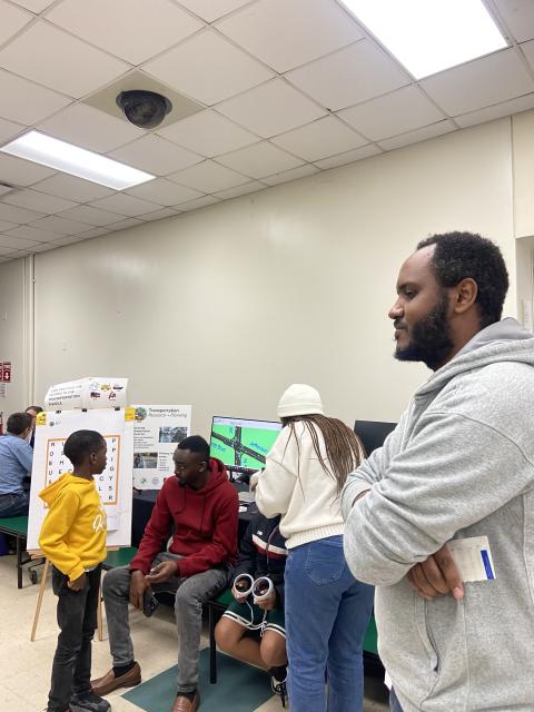Students and adults engage with a transportation planning project display in a classroom setting with posters and a computer monitor.