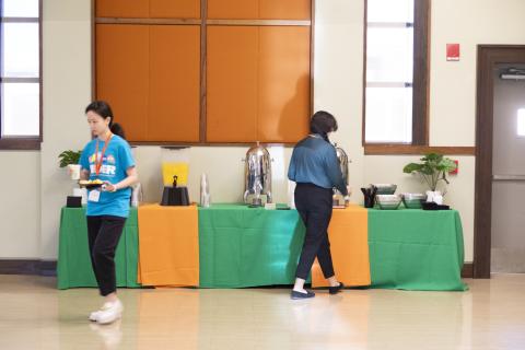 Two volunteers staffing a beverage station with green and orange tablecloths, offering drinks and refreshments at an indoor event.