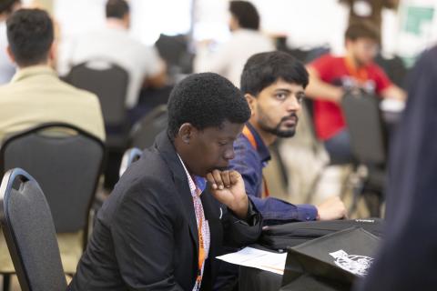 A man wearing a lanyard and blazer sits at a table during a conference or event, reviewing documents while other attendees stand and interact in the background.