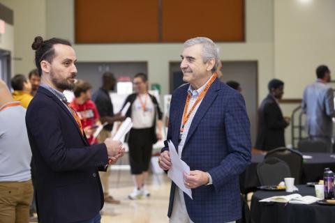 Two men wearing orange lanyards converse at a conference or academic event in a large indoor venue with other attendees visible in the background.
