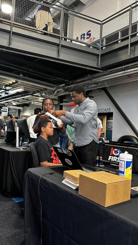A mentor adjusts a participant's headwear at a FIRST Robotics event in an indoor facility with industrial ceiling and black tables.
