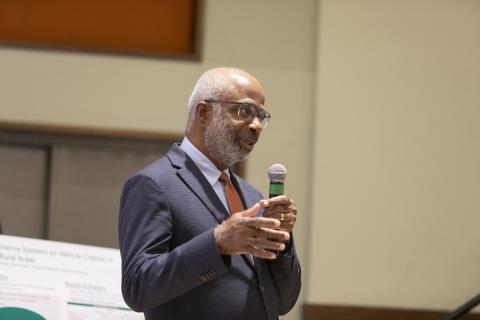 A man in a dark blazer and orange tie holds a microphone while speaking in an indoor presentation setting with beige walls.
