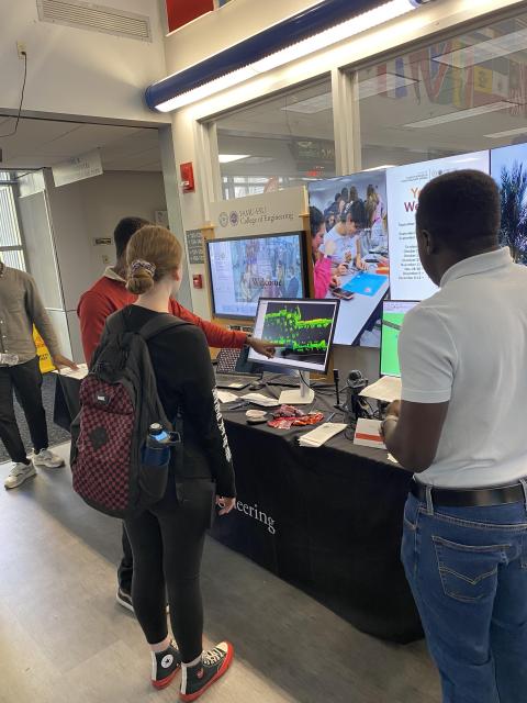 Students and visitors interact with displays at an engineering outreach event in a university lab, with monitors showing research projects and promotional materials on the table.