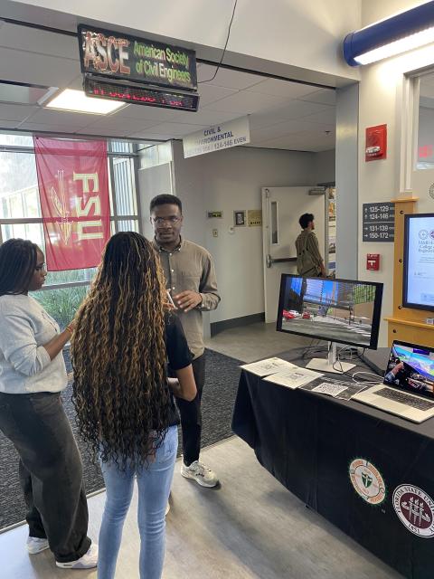 A male researcher presents civil engineering project findings to two female students in a university lab, with monitors displaying architectural renderings and an ASCE banner visible overhead.
