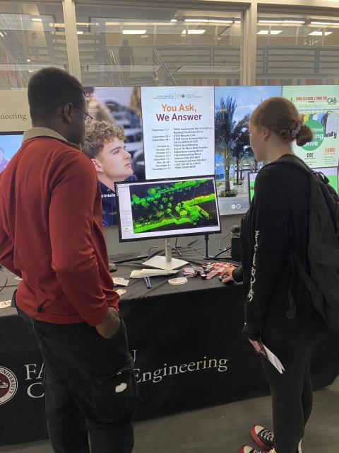 Two students examine a green architectural rendering displayed on a monitor at a university engineering department booth during an event.