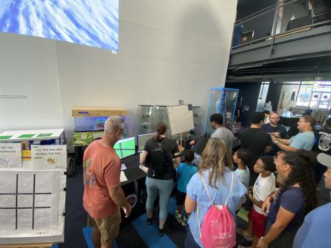 A group of visitors gathered around interactive displays and gaming stations in a research lab or science center, with a presenter demonstrating educational content on screens.