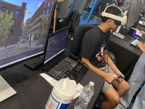 Student wearing a VR headset works at a computer workstation displaying a 3D urban environment, with multiple monitors and research equipment visible.