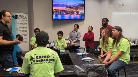 Camp Challenger participants in green t-shirts gather around a table during a presentation at the Northwest Regional Data Center, with an adult speaker gesturing to the group.