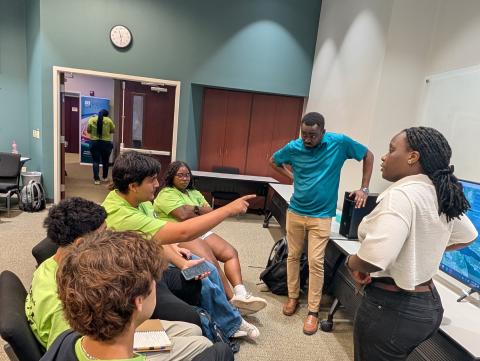 A group of students in neon yellow shirts sit and stand in a circle during an indoor discussion or workshop, with an adult facilitator in a turquoise shirt engaging with the group in a campus room.