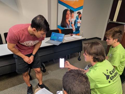 An adult instructor in a maroon shirt demonstrates technology to four students in bright green shirts in an indoor classroom setting, with a laptop and mobile devices visible.