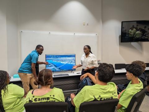 Two instructors present to a group of students wearing lime green camp shirts in a classroom with a monitor displaying an aerial map view.