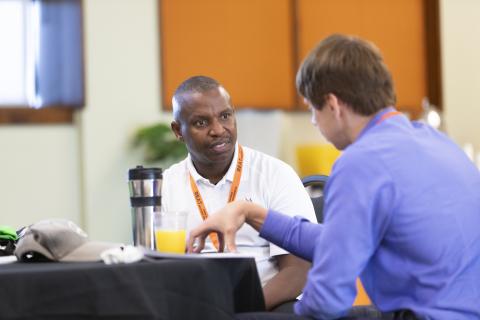 Two men having a conversation at a table during what appears to be a conference or networking event, with orange and white walls visible in the background.