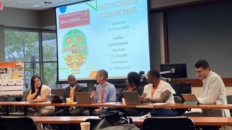 Six panelists seated at a conference table during an Annual UTE Conference panel discussion on practitioners and academic topics, with a projection screen displaying panelist names and moderator information.