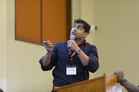 Man wearing navy shirt and ID badge speaking at podium with microphone in academic setting.