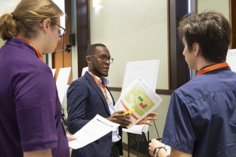 A man in a dark blazer holding a colorful diagram discusses research with two colleagues wearing orange lanyards in an indoor setting.