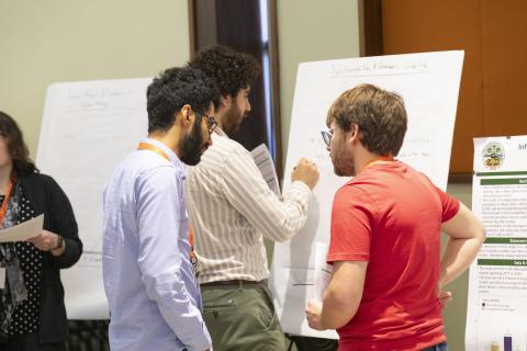 Three researchers discuss findings at a poster presentation session, with whiteboards displaying research content visible in the background.