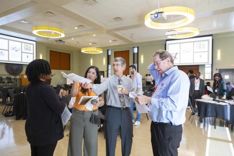 Professionals and attendees engage in discussion at a conference or workshop event in a modern meeting room with yellow pendant lighting.