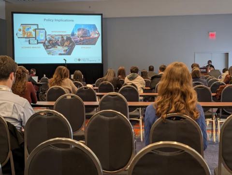 Audience members seated in an auditorium watching a presentation slide titled "Policy Implications" displayed on a large screen, showing intervention scenarios and efficiency analysis.