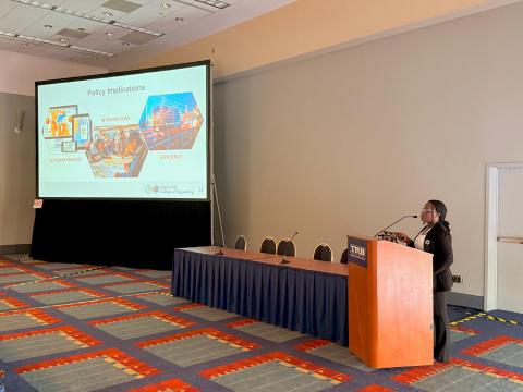Speaker presenting at podium during academic conference with projection screen displaying "Policy Implications" slide from FAMU-FSU College of Engineering presentation.