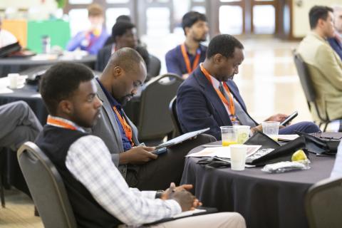 Attendees wearing orange lanyards sit at a table during a conference or workshop, reviewing materials and taking notes.