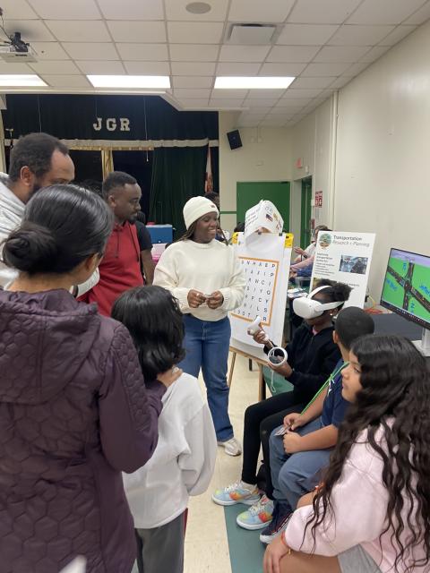 Student presenters demonstrate transportation research project with VR headset and poster board to audience in school gymnasium.