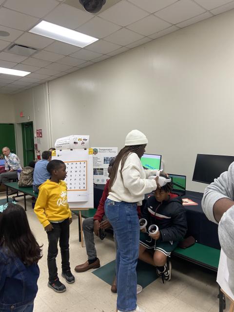 A researcher wearing a white hard hat demonstrates a transportation planning project to students gathered around a table with laptops and a word puzzle display in a classroom setting.