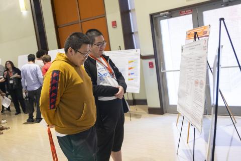 Two students present research posters at an academic conference, with visitors examining displays on easels in a university hallway.