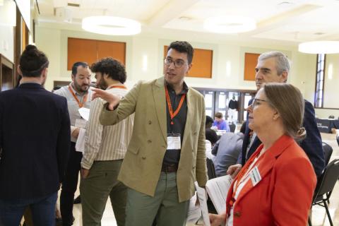 Attendees at a conference or networking event wearing ID badges engage in conversation in a bright indoor space with orange accents and round ceiling lights.