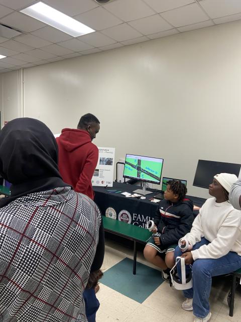 Community members gather around a table displaying a digital transportation planning project on monitors, with presenters in red and black hoodies explaining the research.