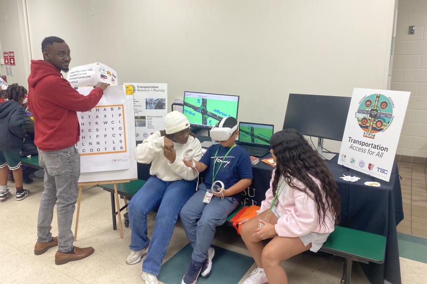 Students engage with a transportation research exhibit featuring interactive displays, a VR headset demonstration, and educational posters about transportation accessibility and planning.