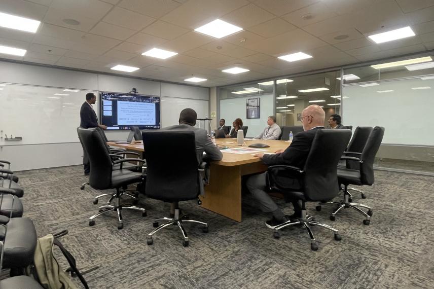 Business professionals in a conference room gathered around a wooden table with one person standing at a display screen presenting data.