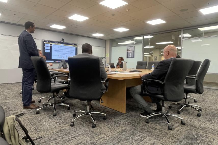 Researchers gathered around a conference table in a modern office meeting room, with one person standing and presenting on a large wall-mounted display screen.