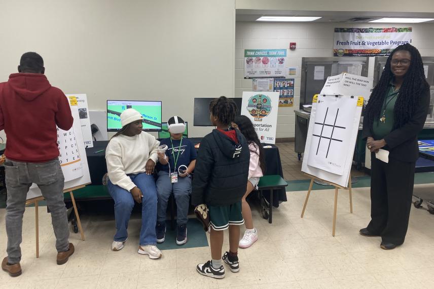 Students and staff participate in an interactive educational activity in a school cafeteria, with game boards and informational posters displayed on easels.
