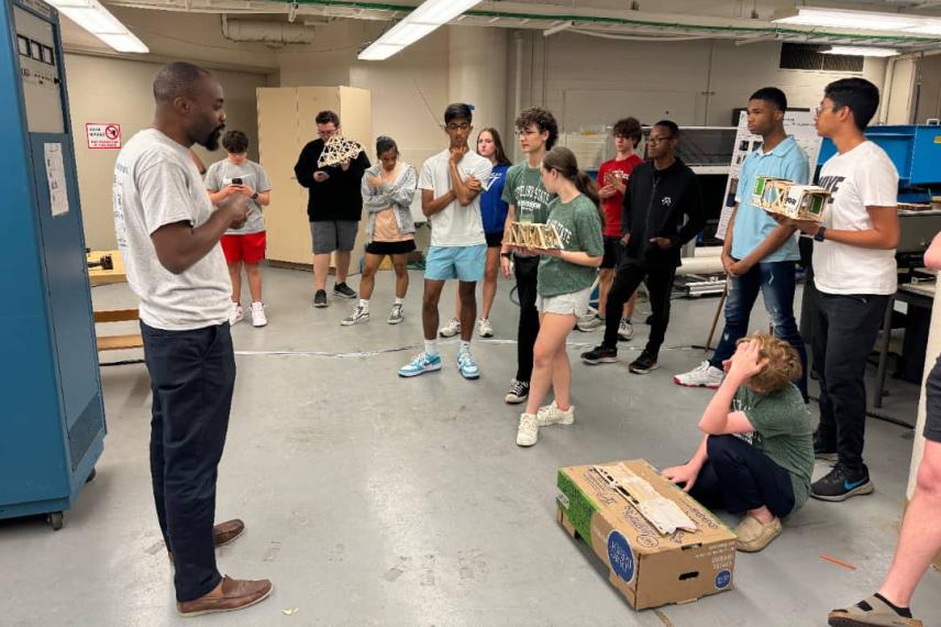 An instructor leads a group of diverse young people gathered in a laboratory or workshop space, demonstrating or discussing materials near a cardboard box on the floor.