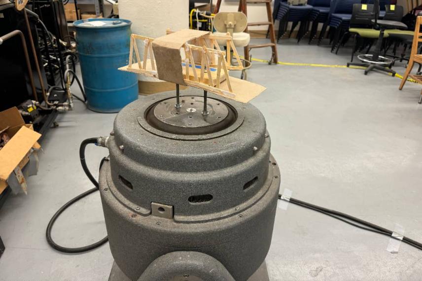 A wooden model airplane sits atop a gray cylindrical motor or turbine in a laboratory workspace, with storage shelves and equipment visible in the background.