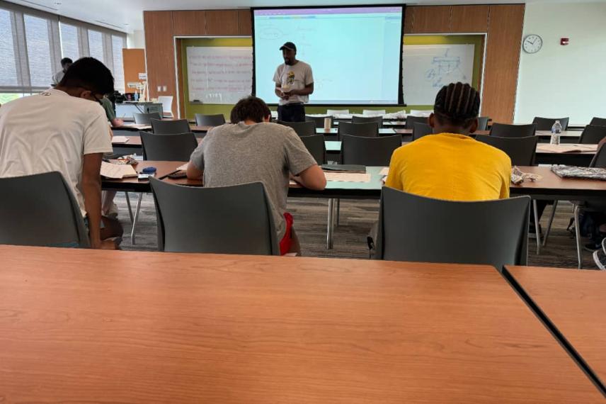 Students seated at desks in a classroom attend a lecture presented on a large projection screen by an instructor standing at the front of the room.