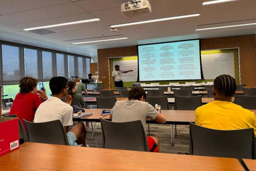 Students seated in a lecture hall watching a presenter standing before a large screen displaying structural bridge diagrams.