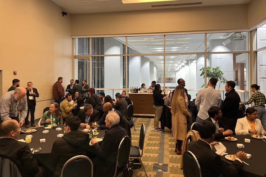 Networking event in a modern conference room with attendees seated at tables and standing, with beverages and refreshments visible.